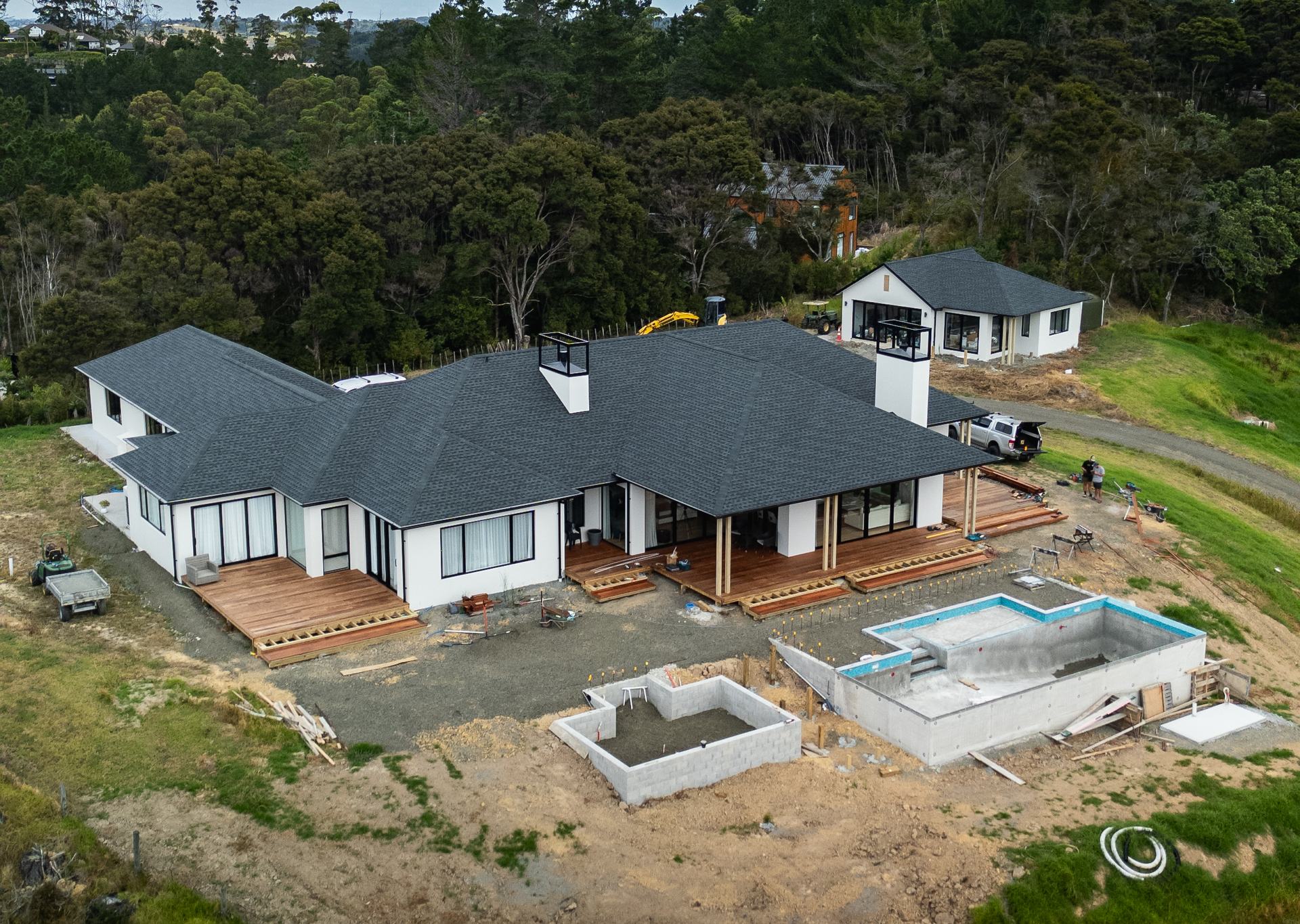 Aerial view of a new white home with a dark roof and timber decks, its concrete pool still being built